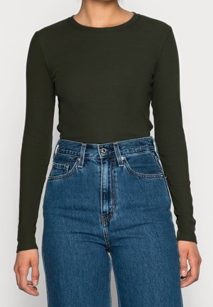 Personne portant un haut moulant à manches longues vert foncé rentré dans un jean en denim bleu taille haute avec poches avant et passants de ceinture.