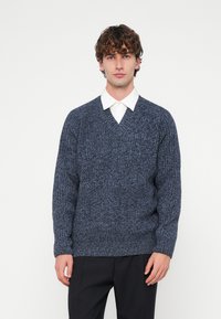 Tumši zils adīts džemperis ar V veida izgriezumu, teksturētu audumu un garām piedurknēm. Valkāts virs baltas krekla ar apkakli, kopā ar tumšām biksēm.