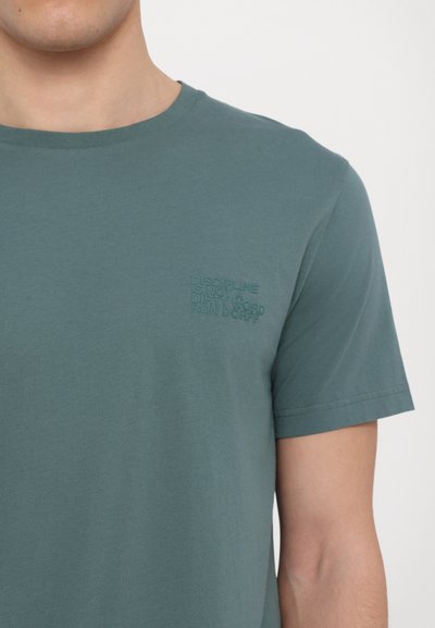 T-shirt en coton turquoise avec un col rond. Il présente un texte brodé subtil sur la poitrine gauche, mettant en valeur des phrases dans des couleurs contrastées.