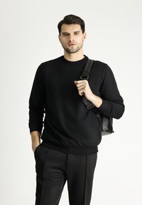 Maglione nero a collo rotondo realizzato in tessuto morbido, con polsini e orlo a costine, indossato con pantaloni neri su misura e una borsa a tracolla nera.