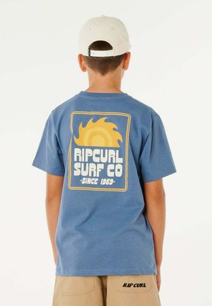Junge trägt ein blaues Rip Curl Surf Co. T-Shirt mit gelbem Sonnenlogo und beige Shorts mit Rip Curl Schriftzug, von hinten gesehen.