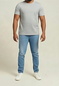 Homme portant un t-shirt à manches courtes gris clair, un jean bleu clair et des baskets blanches, debout devant un fond beige uni.