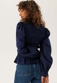 Vrouw met krullend haar, gekleed in een marineblauwe blouse met plooien en pofmouwen, en een blauwe spijkerbroek, zijwaarts kijkend tegen een effen achtergrond.