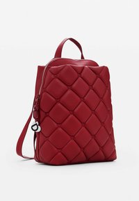 Mochila de cuero acolchado rojo con un asa superior, correa de hombro ajustable y cierre de cremallera. Cuenta con una etiqueta circular adjunta a la cremallera.