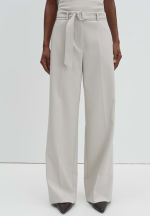 Beige weite Hose mit hohem Bund, Gürtelschlaufen und passendem Stoffgürtel, getragen mit spitzen dunkelbraunen Schuhen.