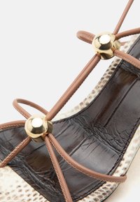 Καφέ σανδάλι με λουράκια, διακοσμημένο με χρυσές σφαιρικές λεπτομέρειες και ανάγλυφη εσωτερική σόλα σε μαύρο και μπεζ χρώμα.