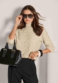 Frau mit langen braunen Haaren, die einen beigen strukturierten Rollkragenpullover, schwarze Hose, große Sonnenbrille, schwarze Handtasche und zwei Armreifen trägt.