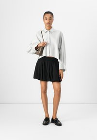Chemise blanche à rayures, jupe évasée noire, mocassins noirs, tenant un petit sac marron, sur un fond neutre.