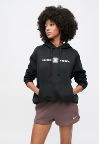 Fiatal nő göndör hajjal, fekete Nike Shox kapucnis pulóvert és mályva színű Nike rövidnadrágot visel, kezét a kapucnis pulóver zsebében tartva, oldalra nézve.