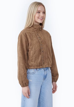 Kvinde med langt blondt hår bærer en brun ruskindsjakke med metaløjer og lyseblå højtaljede jeans, står og smiler.