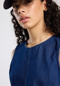 Denimmekko, jossa on pyöreä kaula-aukko ja olkaimet, sinisessä sävyssä, näkyvät saumat ja hieno rakenne. Päällä kultaketju.