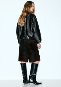 Schwarze übergroße Lederjacke, braune Wildledershorts und kniehohe schwarze Lederstiefel mit spitzen Zehen. Modell von hinten gesehen.