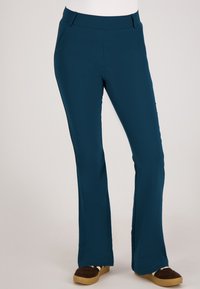 Teal flared broeken van rekbaar materiaal met voorkruiszakken, riemlussen en een gladde textuur. Draagt met bruine, suèdeachtige schoenen.