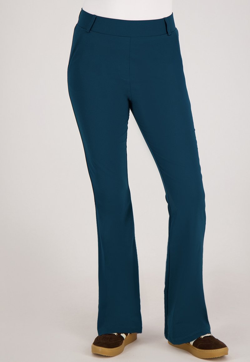 Teal flared broeken van rekbaar materiaal met voorkruiszakken, riemlussen en een gladde textuur. Draagt met bruine, suèdeachtige schoenen.