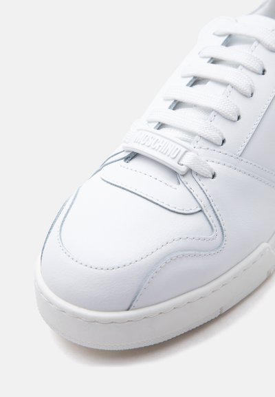 Witte leren sneaker met witte veters, gedetailleerde stiksels en een merklabel aan de veter met de tekst "MOSCHINO" op een gladde zool.