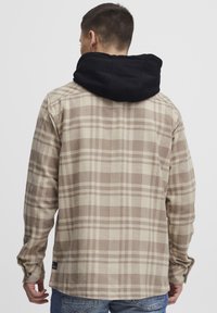 Bruine en beige geruite flanellen shirt met een zwarte capuchon. Heeft lange mouwen en een gebogen zoom, gemaakt van een gestructureerde stof.
