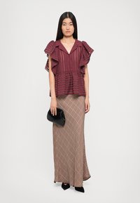 Bordeaux peplumtop met gerimpelde mouwen, gecombineerd met een lange, gemusterde maxirok in beige en bruin. Zwarte handtas met een gestructureerde afwerking.