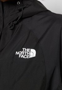 Czarna kurtka wykonana z wodoodpornej tkaniny z kapturem. Charakteryzuje się wyraźnym białym logo na klatce piersiowej oraz gładką teksturą.