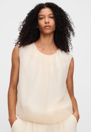 Vrouw met krullend haar die een mouwloze lichtbeige blouse draagt met haar handen in de zakken van een bijpassende broek, kijkt recht vooruit tegen een effen achtergrond.