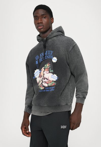 Young man wearing a faded black hoodie with a colorful graphic and text, paired with dark pants, standing against a plain background.
