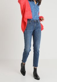 Donkerblauwe spijkerbroek met hoge taille, gecombineerd met een lichtblauw overhemd met knopen. Een rode cardigan hangt over de schouders. Zwarte enkellaarsjes.