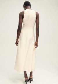 Kermavärinen hihaton mekko, jossa on tiukka yläosa ja liehuva, nilkkapituinen hame. Ominaisuuksiin kuuluu siisti muotoilu ja karkea helman yksityiskohta.