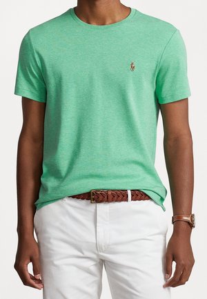 Mann trägt ein grünes kurzärmliges T-Shirt mit einem kleinen gestickten Logo, weiße Hose, braunen geflochtenen Gürtel und braune Lederarmbanduhr.