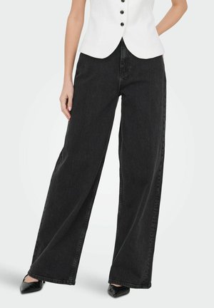 Kvinde iført sorte vidde bukser, hvid knappet vest og sorte spidse hæle, stående med hænderne nær lommerne.