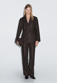 Žena stojící v tmavě hnědém dvouřadém saku, se stejnobarevnými kalhotami s širokými nohavicemi, držící hnědou semišovou kabelku.