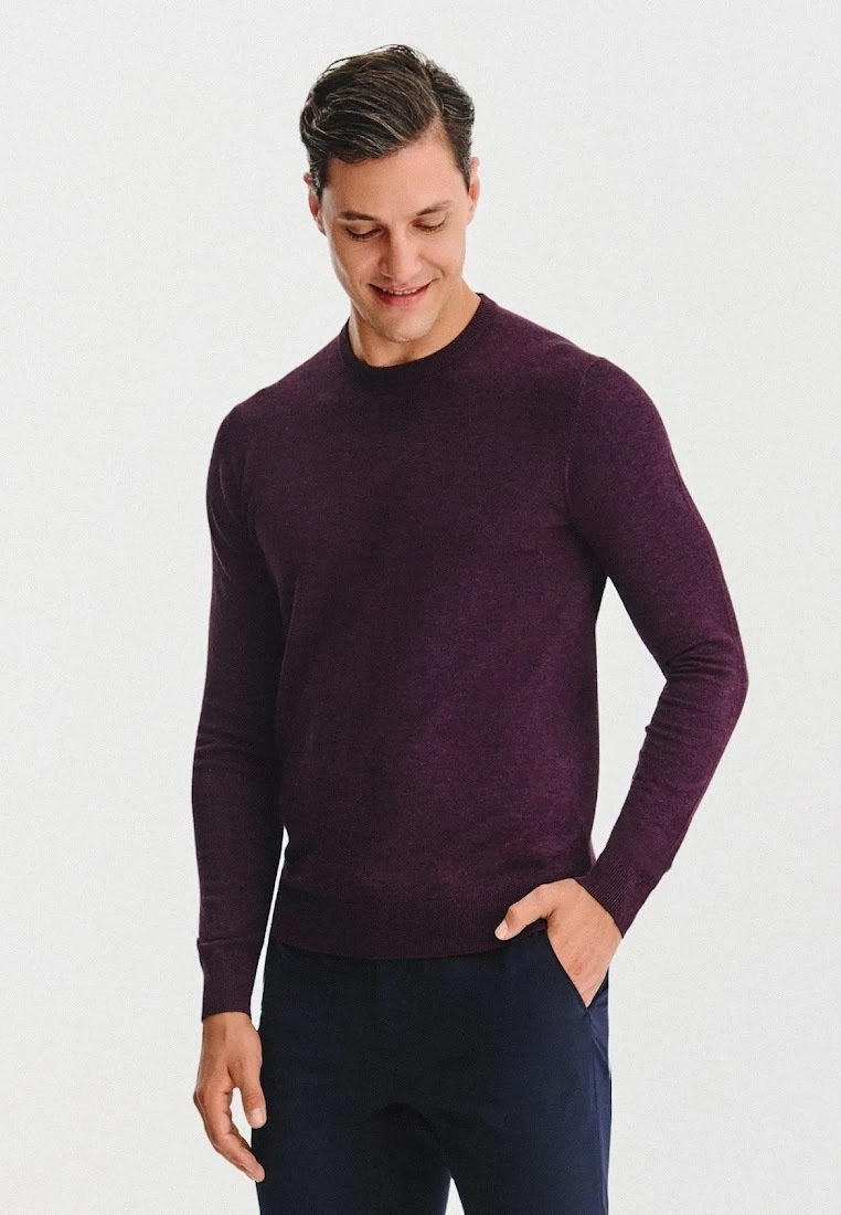 Mężczyzna w dopasowanym, ciemnofioletowym swetrze z długim rękawem i ciemnych spodniach, patrzący w dół, z jedną ręką w kieszeni, na jednolitym tle.