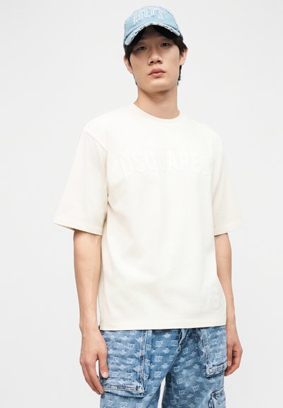Jeune homme portant un T-shirt beige clair Dsquared2, une casquette de baseball bleu clair délavée et un pantalon cargo en denim bleu à motifs sur fond blanc.