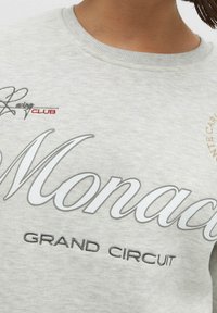 Sudadera gris claro con cuello redondo. Presenta un gran texto "Monaco GRAND CIRCUIT" en blanco y negro, con acentos en rojo y dorado. Textura suave.