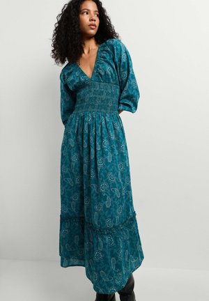 Frau mit lockigem Haar, die ein langes Kleid in Türkis mit Paisley-Muster, Puffärmeln und geraffter Taille trägt, steht vor einer schlichten hellen Wand.