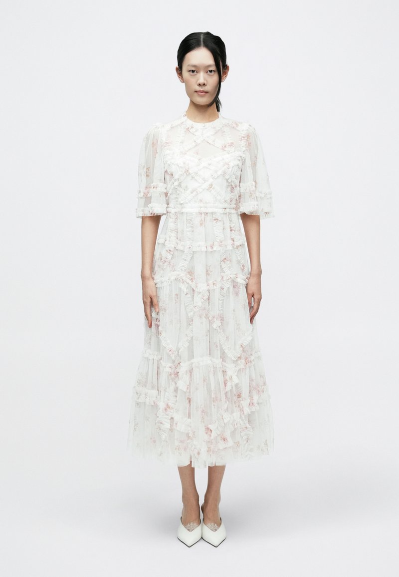 Balta midi kleita ar maigām rozā ziedu rakstām, kurai ir volānu slāņi un uzpūstās piedurknes. Saskaņota ar baltām, smailām kurpēm.