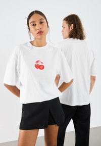 Hvid bomulds T-shirt med et rødt kirsebærgrafik på fronten. Løs pasform med korte ærmer, stylet med sorte shorts.