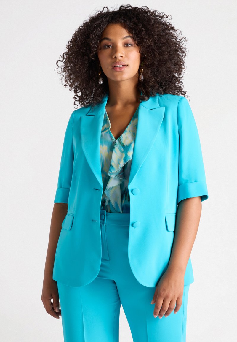 Türkiser Blazer mit kurzen Ärmeln, Reverskragen, doppelten Knöpfen und einer Tasche. Trägt man über einem gemusterten blau-grünen Oberteil.