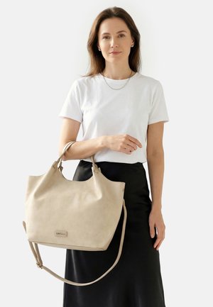Vrouw met lang bruin haar, gekleed in een wit T-shirt en een zwarte rok, die een grote beige draagtas met schouderriem vasthoudt tegen een witte achtergrond.