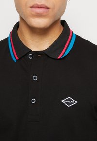 Man in een zwart poloshirt met drie zwarte knopen, een kraag gestreept in blauw en rood, en een wit Replay-logo op de borst.