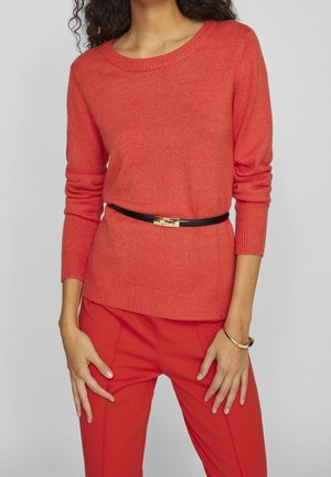 Kobieta w dopasowanym czerwonym swetrze z czarnym paskiem i pasujących czerwonych spodniach, stojąca z rękami przy biodrach na jednolitym tle.