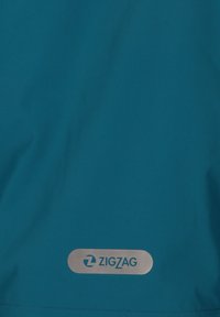 Kobaltblauw waterdicht stof met een zachte textuur, voorzien van een opvallende zilveren logo patch met de naam "ZIGZAG" aan de onderzijde.