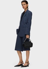 Donna che indossa un blazer blu navy e pantaloni corti coordinate fino al ginocchio, top nero, décolleté neri con tacco a rocchetto, mentre tiene una borsa nera texturizzata con manico in catena.