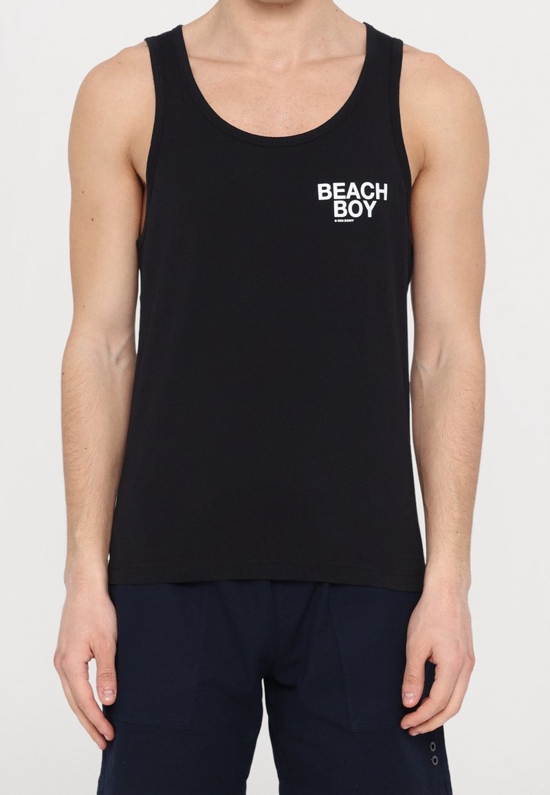 Homme portant un débardeur noir sans manches avec le texte blanc "BEACH BOY" sur le haut à gauche de la poitrine, assorti à un short bleu marine foncé.