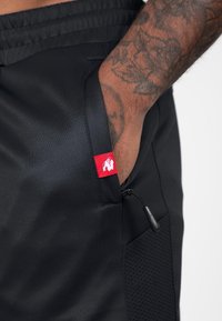 Zwarte sportshorts met een gladde textuur, voorzien van een ritszak, een rode label met een logo en een paneel van gestructureerde stof aan de zijkant.