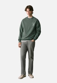 Felpa verde con colletto rotondo e dettaglio del logo, abbinata a jeans grigi e scarpe chiare. Il tessuto sembra morbido con una vestibilità comoda.