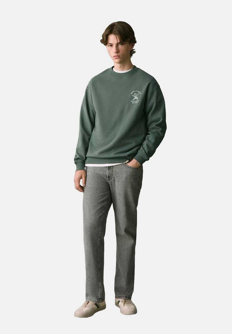 Felpa verde con colletto rotondo e dettaglio del logo, abbinata a jeans grigi e scarpe chiare. Il tessuto sembra morbido con una vestibilità comoda.