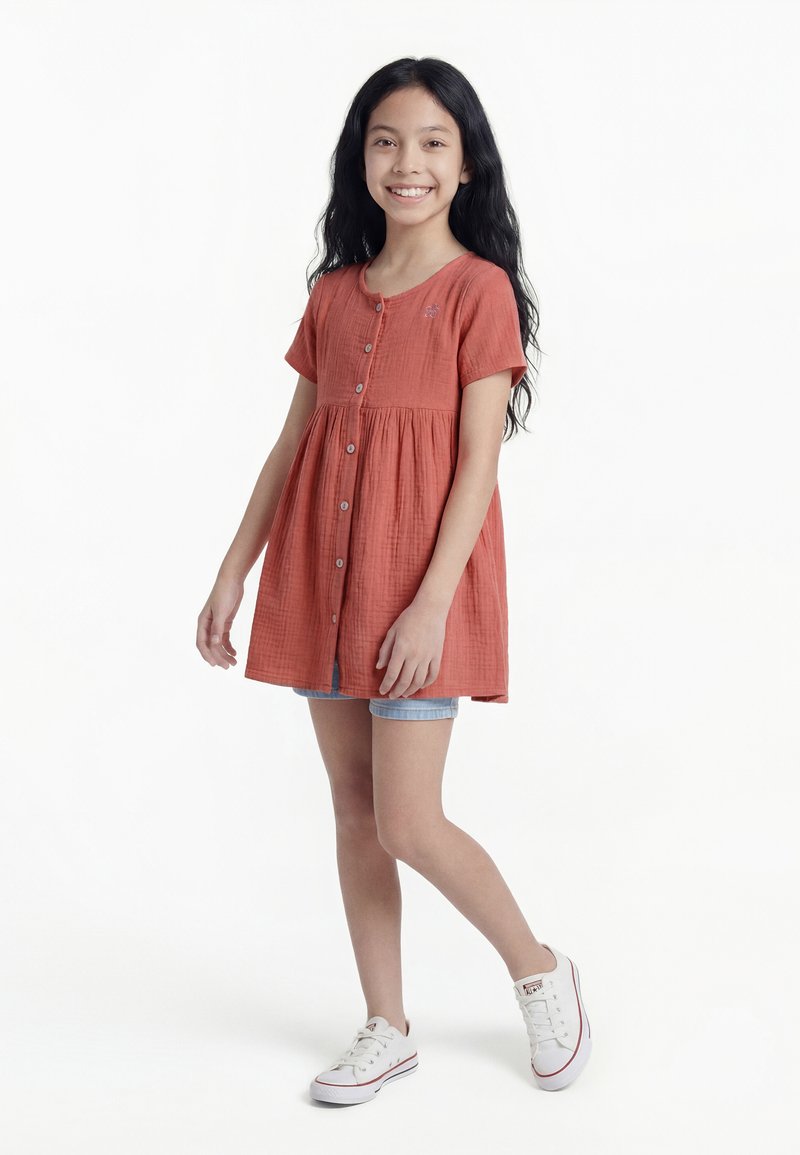 Fille aux longs cheveux noirs, portant une robe couleur rouille boutonnée par-dessus un short en jean et des baskets blanches, souriant et se tenant sur une jambe.