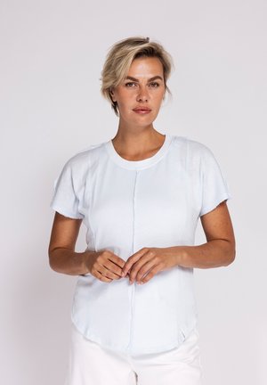 Frau mit kurzen blonden Haaren, die ein weißes kurzärmeliges Oberteil und weiße Hose trägt, steht mit leicht vor dem Körper verschränkten Händen.