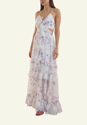 Frau trägt ein weißes Maxikleid im Blumenmuster mit Neckholder, Volants und seitlichen Ausschnitten, kombiniert mit offenen High-Heels.