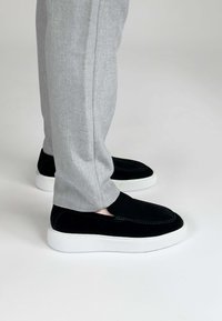 Zwarte suède instappers met een witte rubberen zool, gecombineerd met lichtgrijze, gestructureerde broeken, wat een casual maar eigentijdse uitstraling benadrukt.