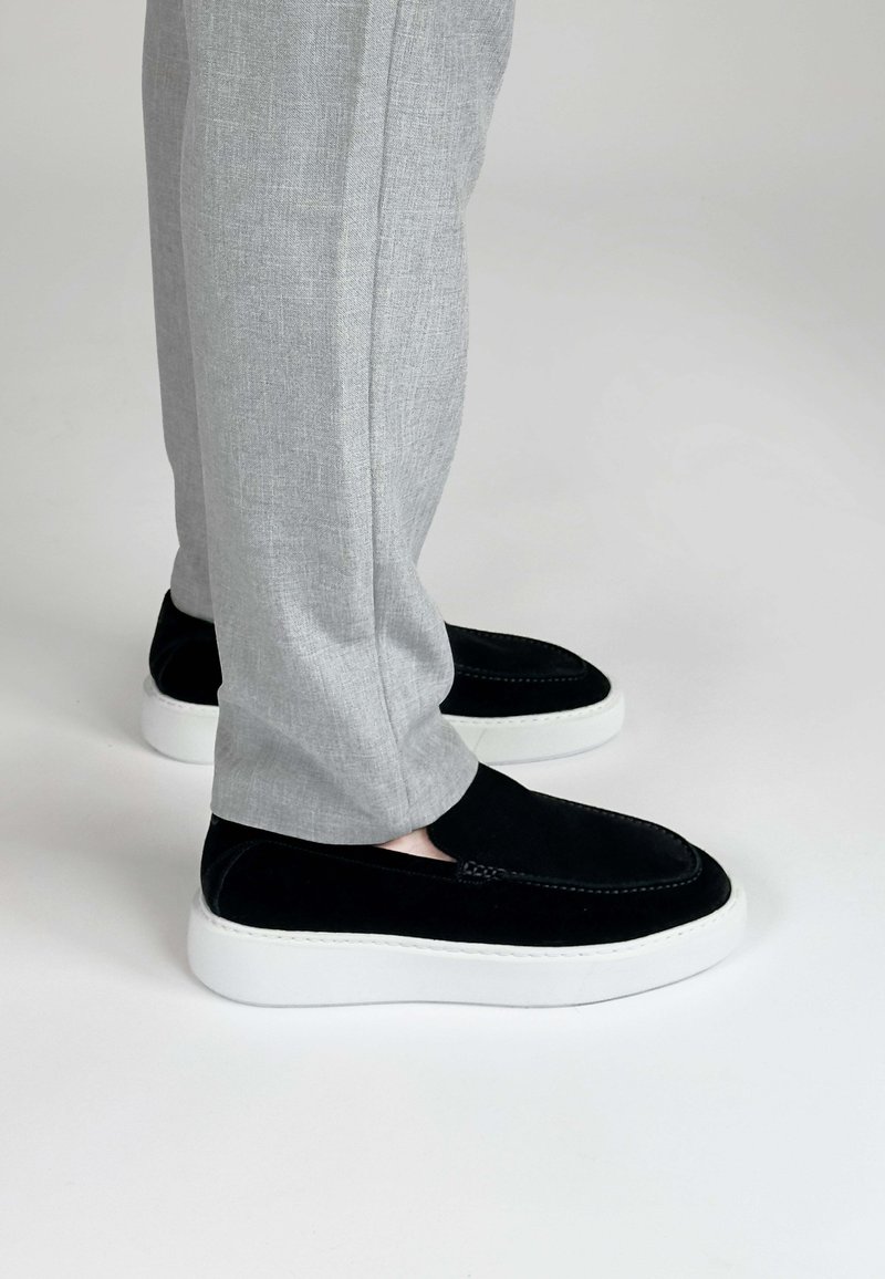 Zwarte suède instappers met een witte rubberen zool, gecombineerd met lichtgrijze, gestructureerde broeken, wat een casual maar eigentijdse uitstraling benadrukt.